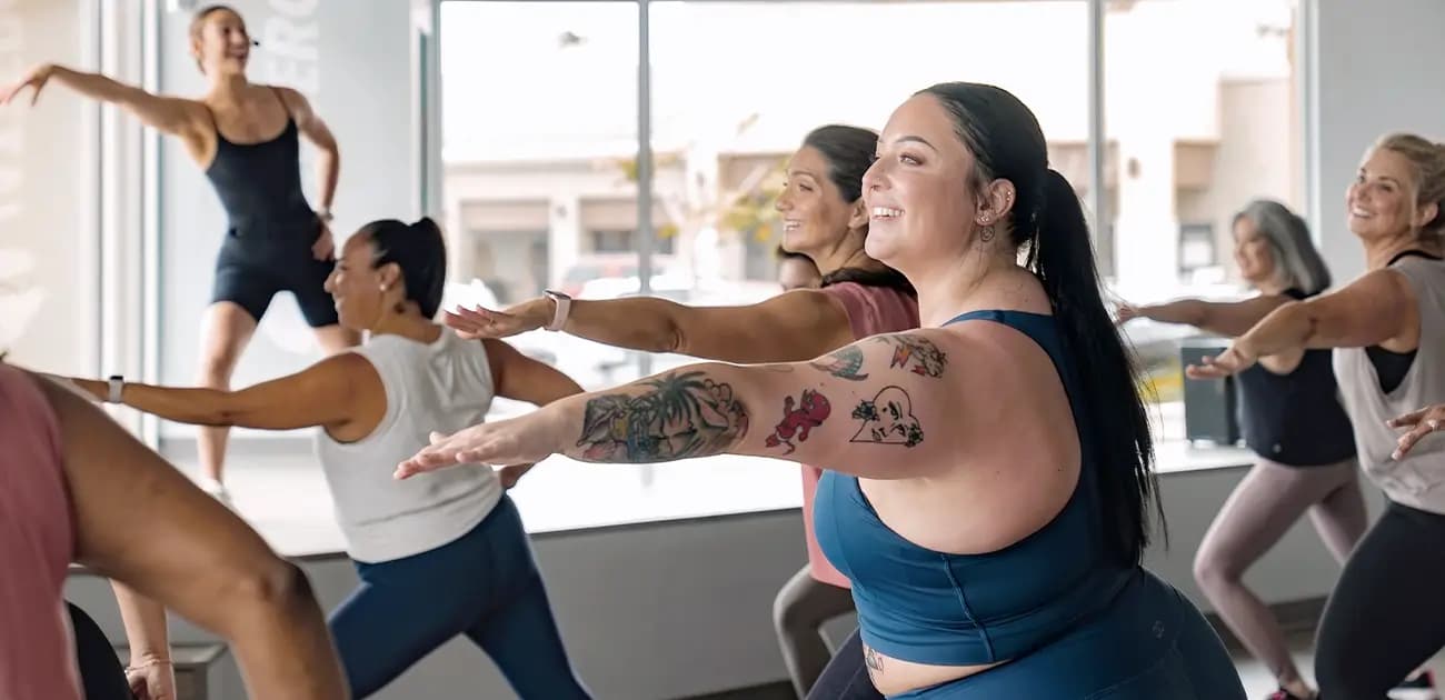 A group of individuals is participating in a lively fitness class, led by an instructor at the front. They are performing a standing exercise with arms outstretched. The room is well-lit with large windows in the background, revealing a view of the outside.