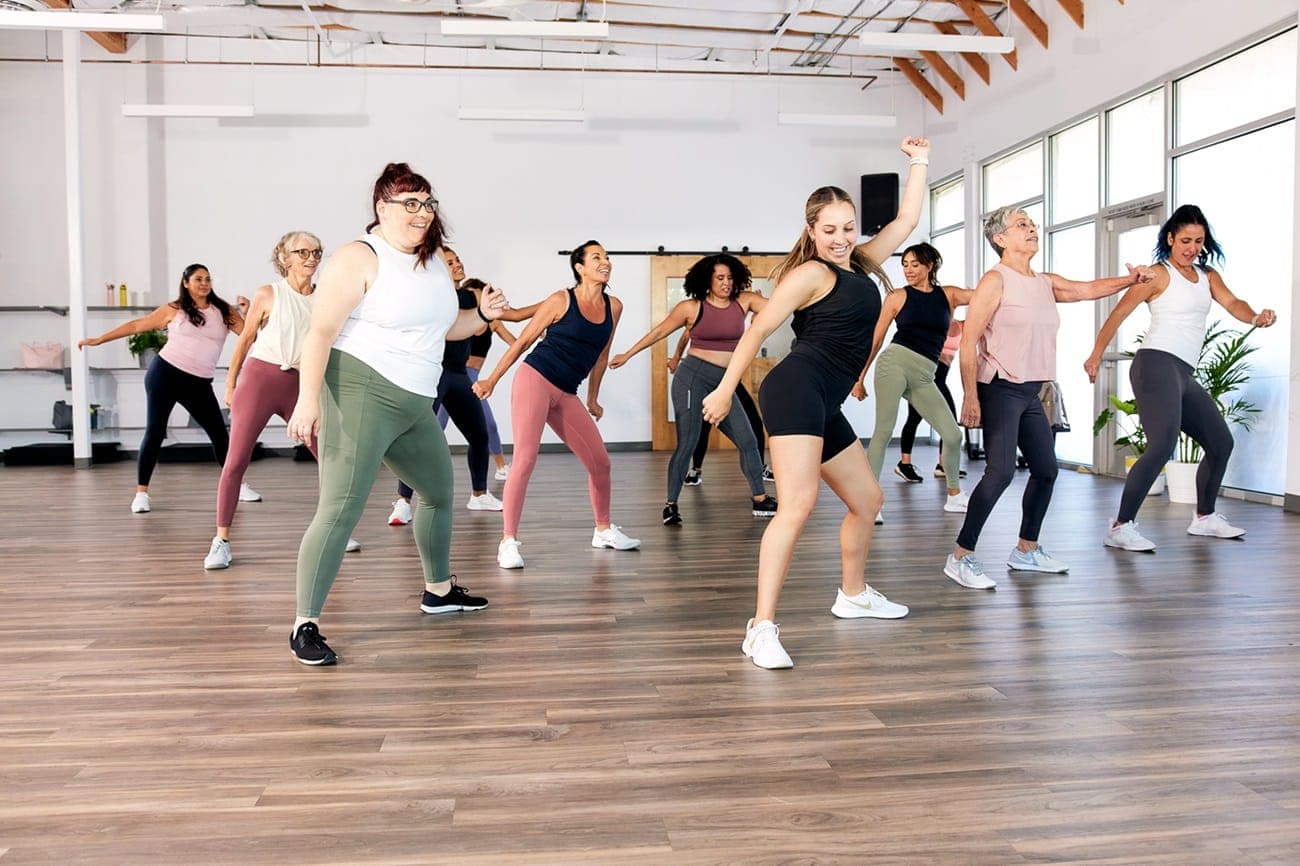 A group of people participate in a dance fitness class in a spacious room with wooden floors and large windows. They are energetically moving in sync, wearing athletic wear in various colors, and appear to be enjoying the workout.