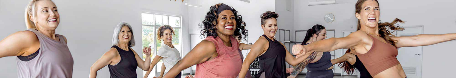 A diverse group of joyful individuals engaged in a dynamic dance or fitness class. They are smiling, energetically moving, and appear to be enjoying themselves. The background shows windows allowing natural light into the room and minimalistic decor.