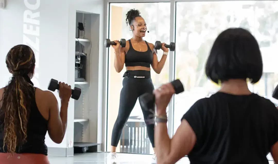 A fitness instructor guides a class in a bright studio. She holds dumbbells while smiling and speaking into a microphone. Participants in the foreground follow her lead, also holding weights. The atmosphere is energetic and focused.