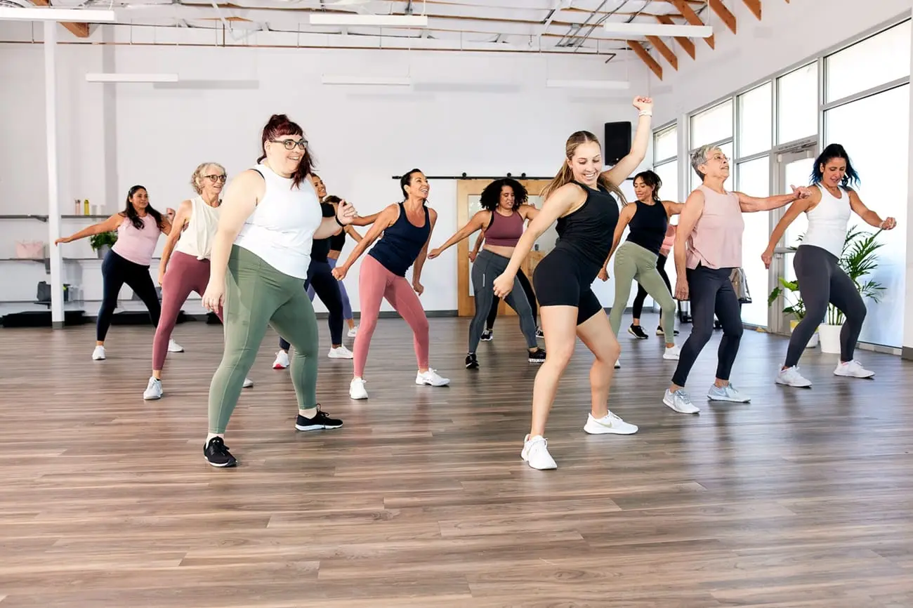 A group of people participate in a dance fitness class in a spacious room with wooden floors and large windows. They are energetically moving in sync, wearing athletic wear in various colors, and appear to be enjoying the workout.